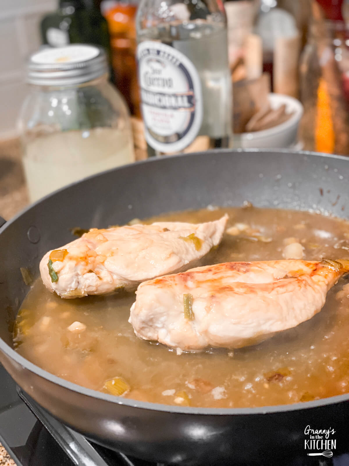 two chicken breasts cooking in a sauté pan, with bottle of tequila in background