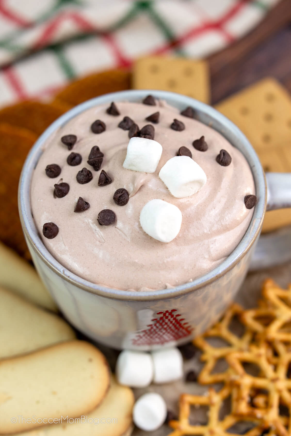 mug of hot cocoa dip surrounded by cookies and pretzels.