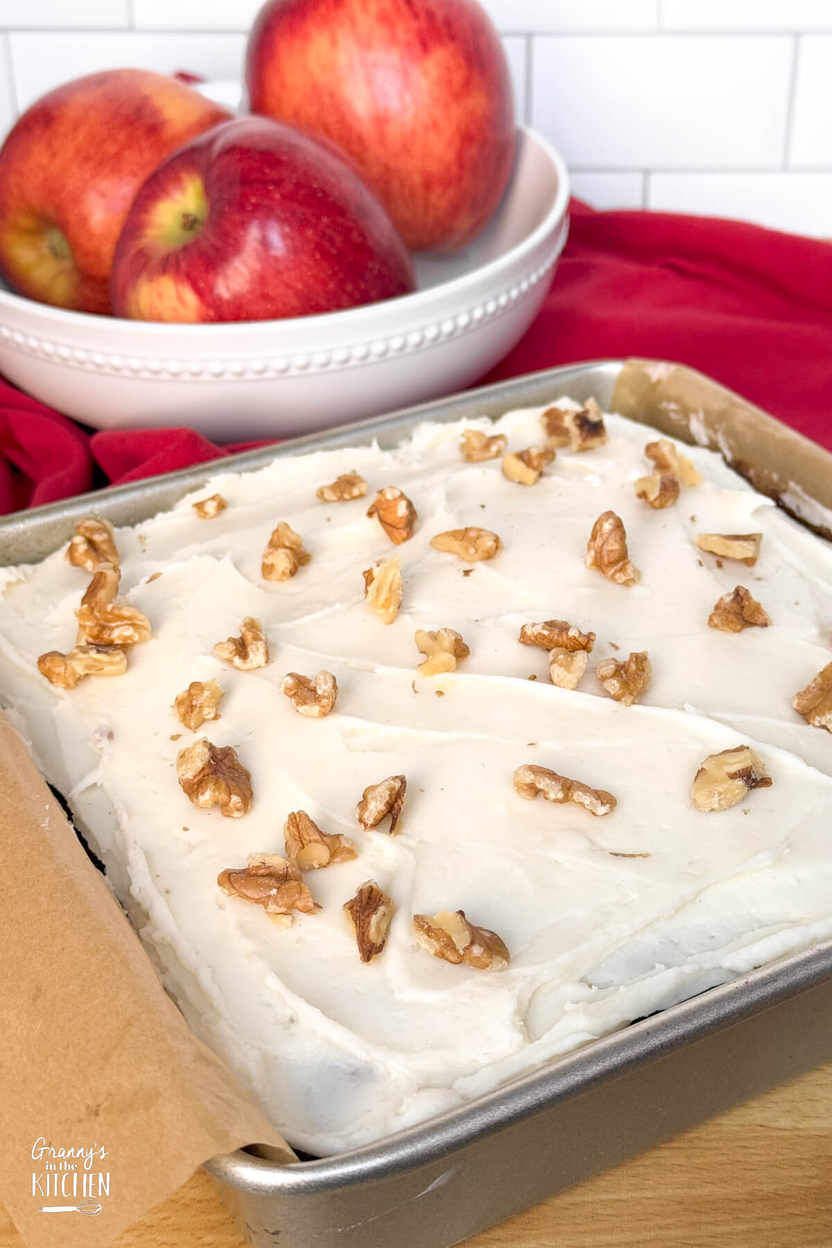 cake in pan with cream cheese icing and chopped walnuts, bowl of apples in background.
