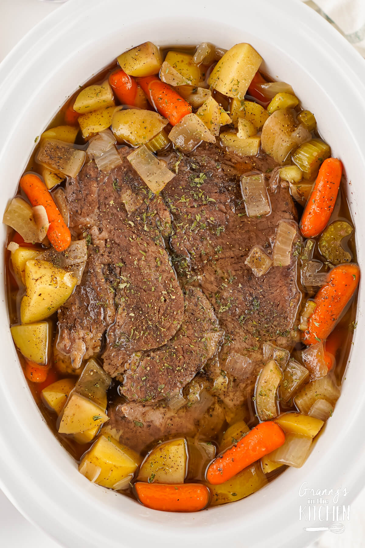 Pot roast with potatoes, onions, celery, and carrots in a white slow cooker.