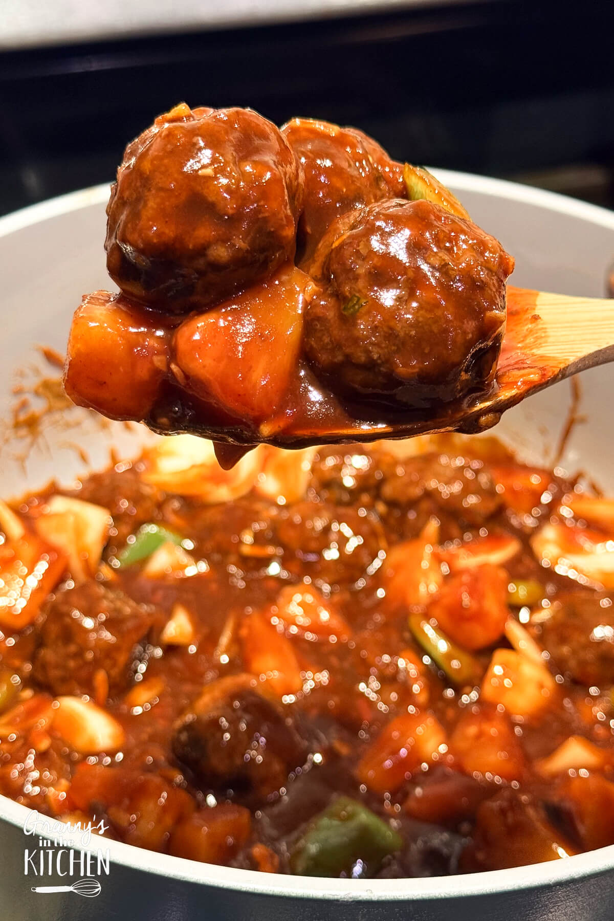 A wooden spoon holding sweet and sour meatballs above a cooking pot.